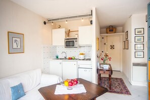 A compact kitchen with a microwave, sink, and cabinets. There's a dining table with a fruit bowl and a small rug.