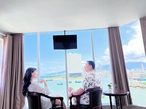A room with a large window offering a city view, a flat-screen TV mounted on the wall, and two chairs facing each other. - Diu Dang Hotel Nha Trang- by Bay Luxury (Bac Nha Trang)