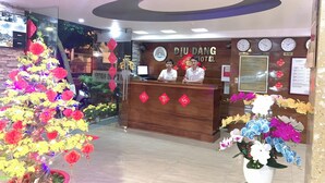 A hotel reception desk with a wooden counter, two receptionists, and a wall clock.