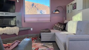 Un salon doté d’une grande fenêtre, d’un téléviseur à écran plat, d’un canapé gris, d’un tapis à motifs et d’un tapis blanc moelleux.
