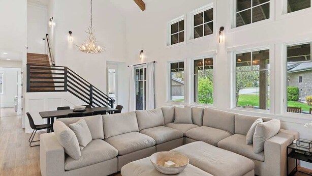 A spacious living room with a large L-shaped sectional sofa, a dining area with a wooden table and chairs, and a staircase with a black railing.