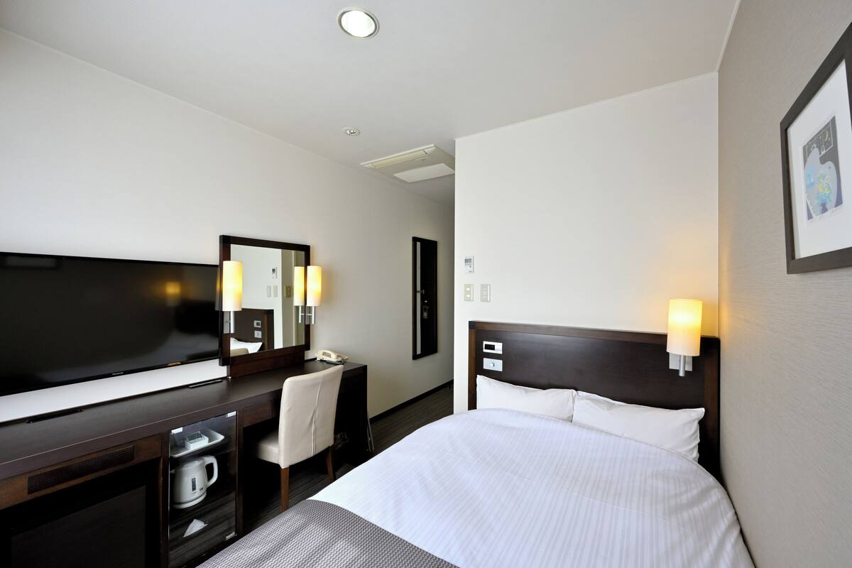 Quarto de hotel com cama, escrivaninha, cadeira, televisão e espelho.