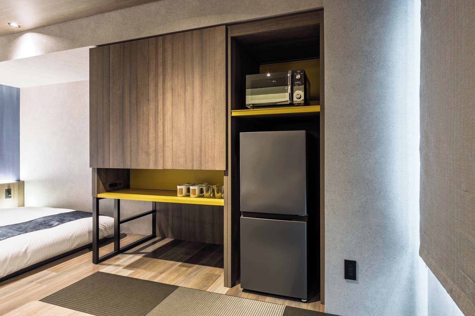 Una habitación de hotel compacta con una cama, una kitchenette integrada con microondas y refrigerador, y un estante pequeño con tazas.