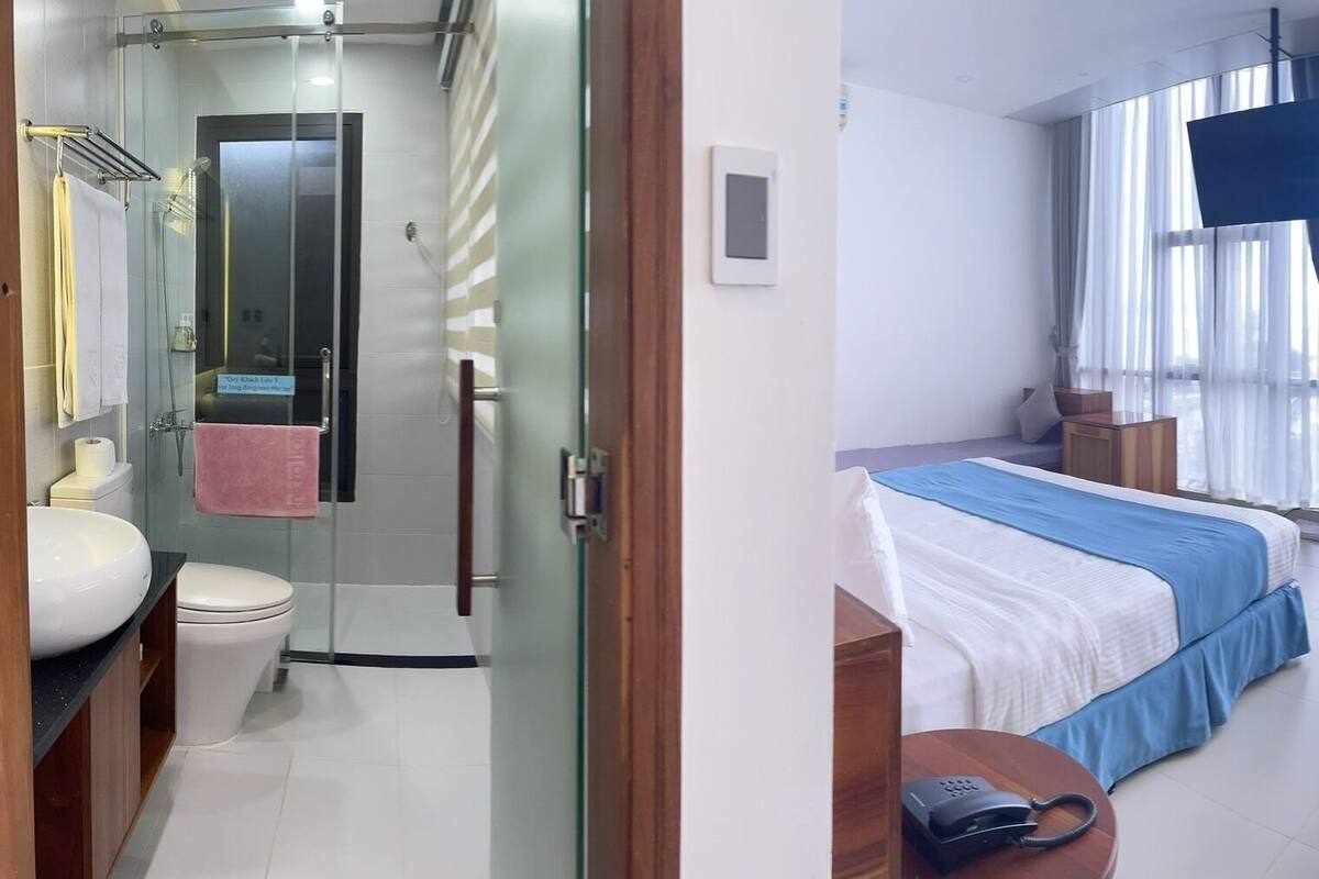 A hotel room with a shower, toilet, sink, bed, bedside table, and a large window with curtains.