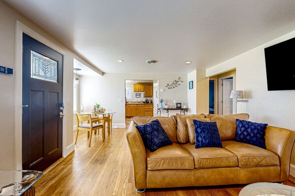 A living room with a brown leather sofa, a dining table with chairs, and a kitchen in the background.