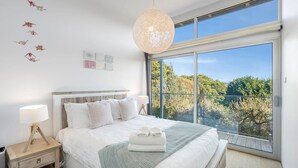 Een slaapkamer met een groot raam, een bed met witte lakens, een nachtkastje met een lamp en een hanglamp.