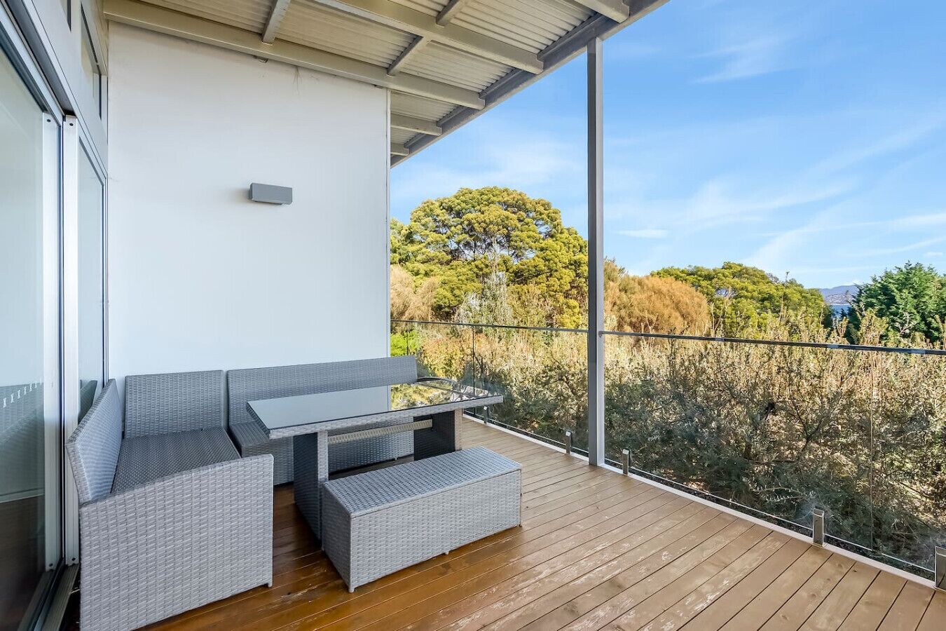 A balcony with wicker furniture and a glass table, overlooking a natural landscape with trees and a clear sky.