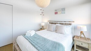 Una cama bien hecha con sábanas blancas y una manta de color azul claro, una lámpara de techo redonda con textura, y una mesita de noche con lámpara.