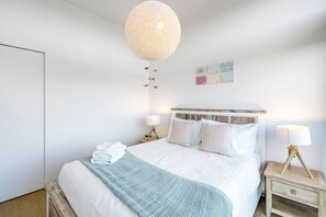 Ein ordentlich bezogenes Bett mit weißen Leinen und einer hellblauen Tagesdecke, eine runde, strukturierte Deckenleuchte sowie ein kleiner Nachttisch mit Lampe.