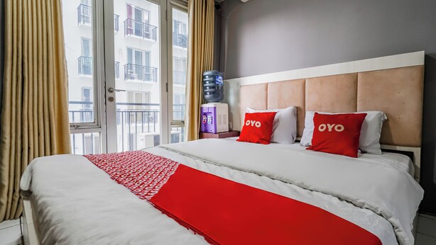 Habitación de hotel con una cama grande, dos almohadas rojas con la marca 'OYO', un dispensador de agua y vista a un edificio desde la ventana.