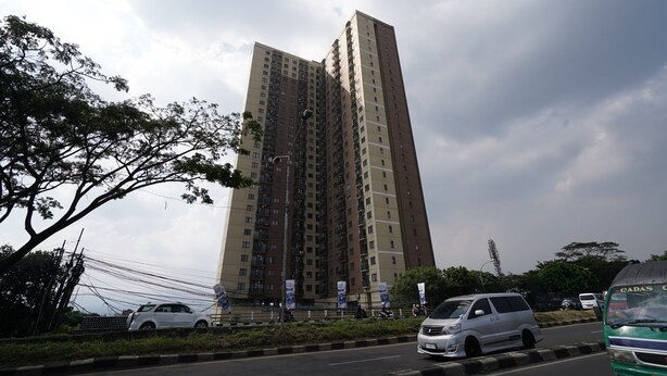 Un edificio residencial alto con varios pisos, rodeado de árboles y vehículos estacionados en la calle adyacente.