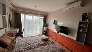 Habitación de hotel con cama, televisor montado en la pared, ventana con cortinas y una pequeña mesa con una silla.