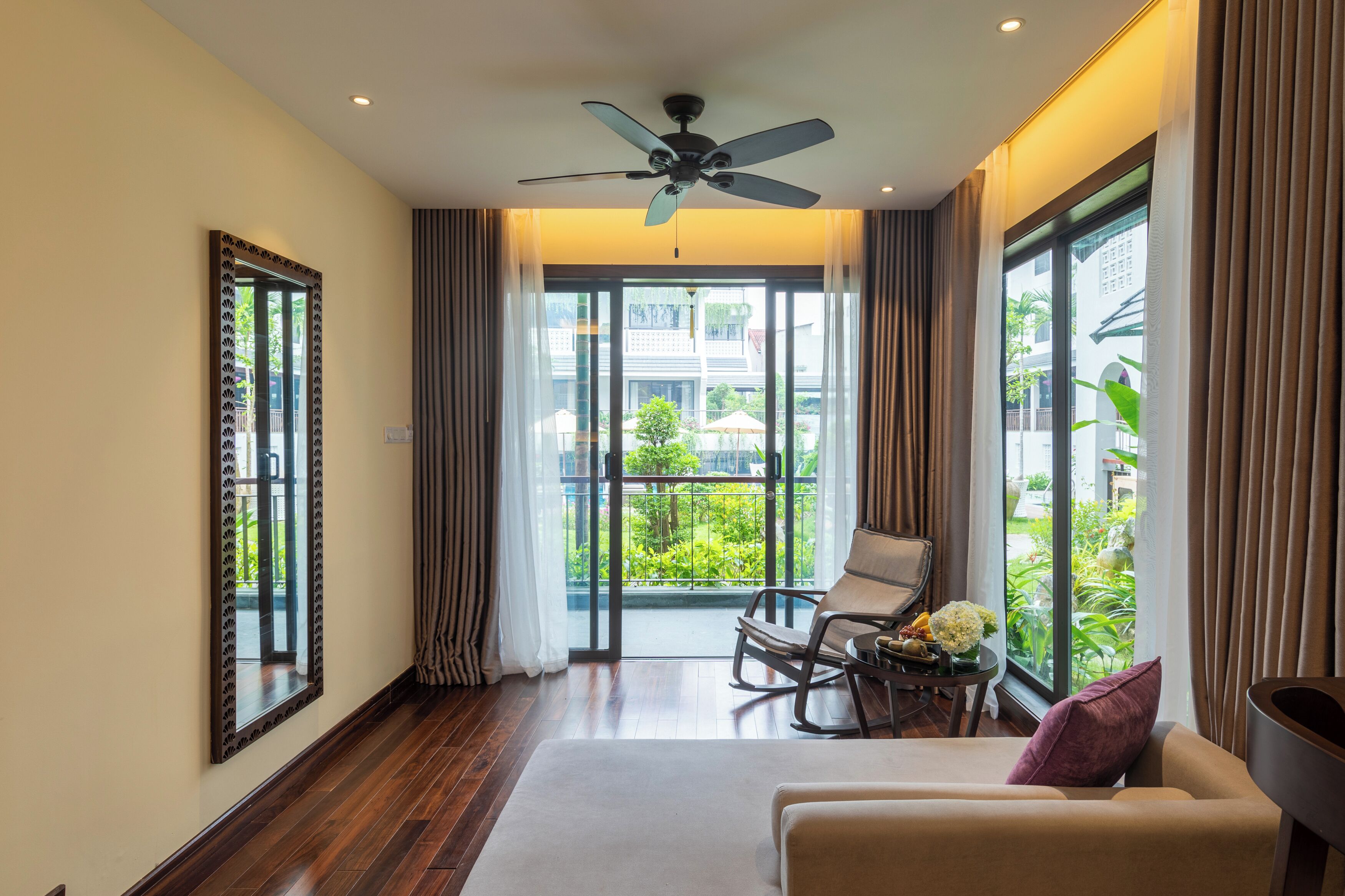 A modern hotel room with a large balcony, a ceiling fan, and a seating area with a table and flowers.