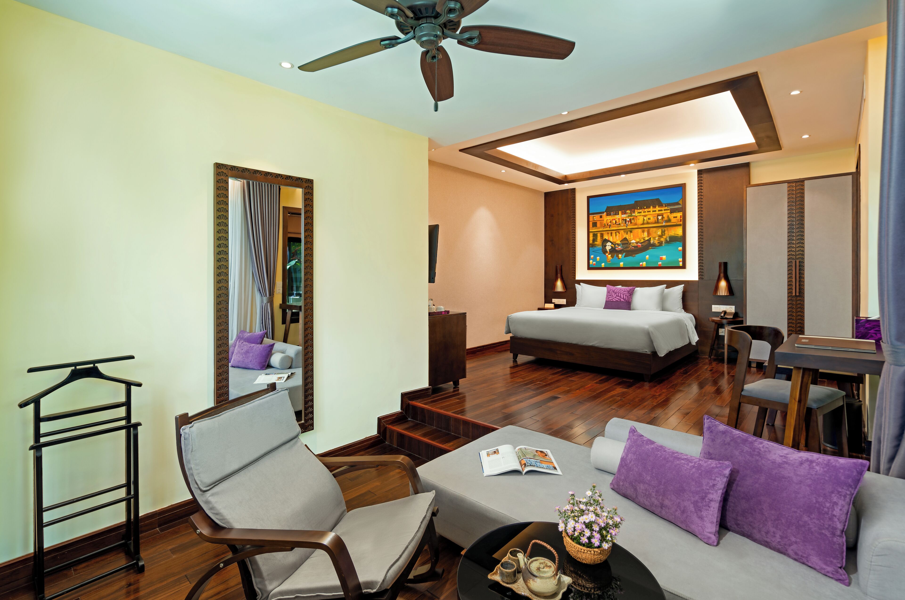 A hotel room with a bed, a sofa, a dining table, a chair, a coffee table, a ceiling fan, and a painting on the wall.