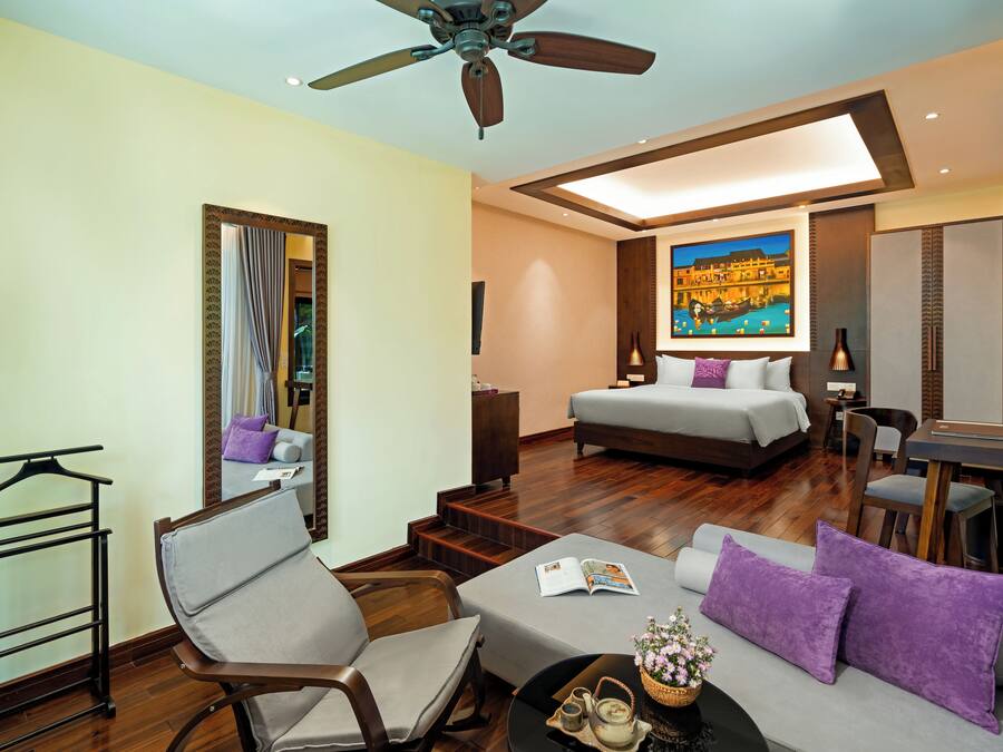A hotel room with a bed, a sofa, a dining table, a chair, a coffee table, a ceiling fan, and a painting on the wall.