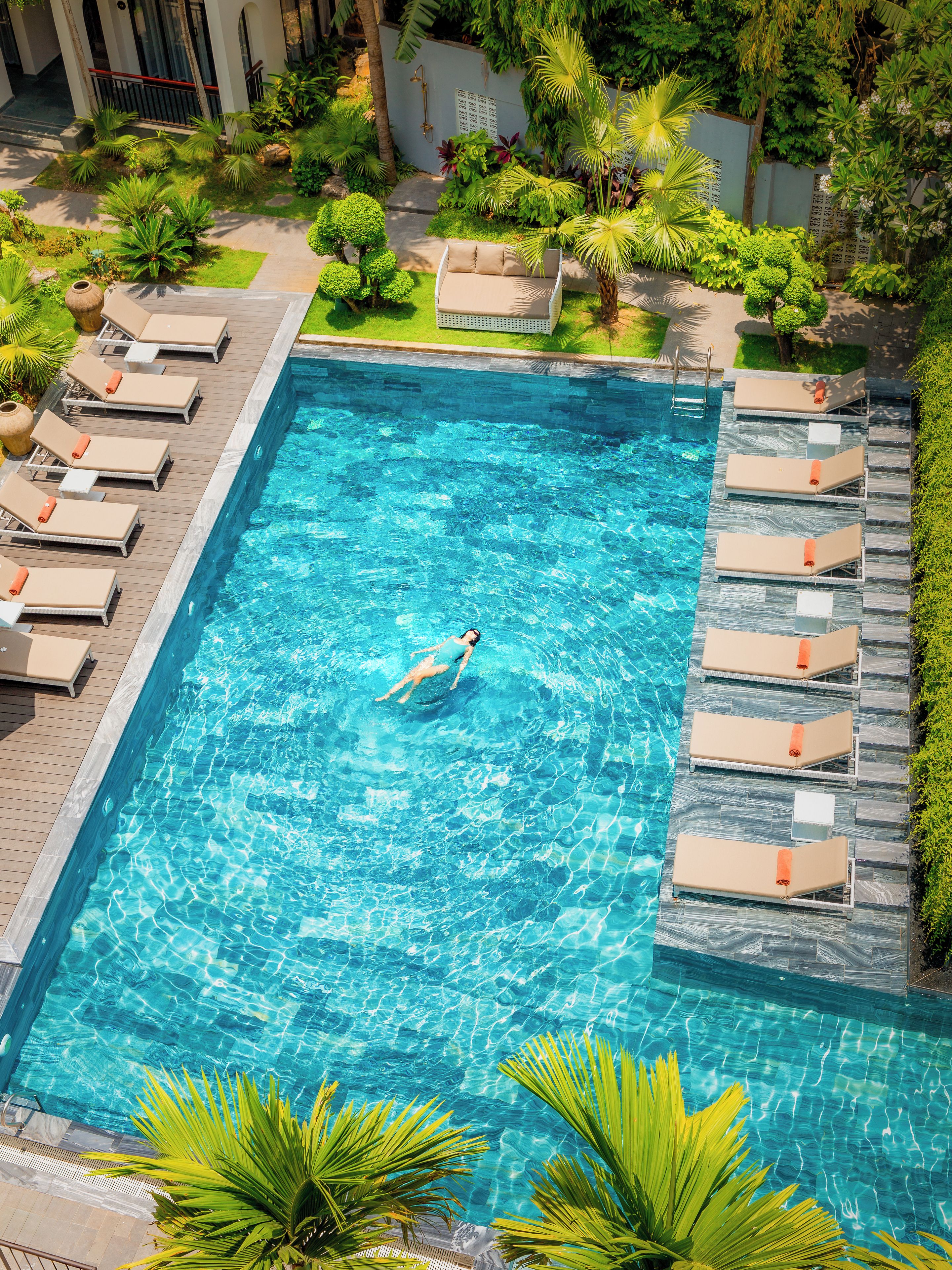 A swimming pool with a person swimming, surrounded by lounge chairs and greenery.