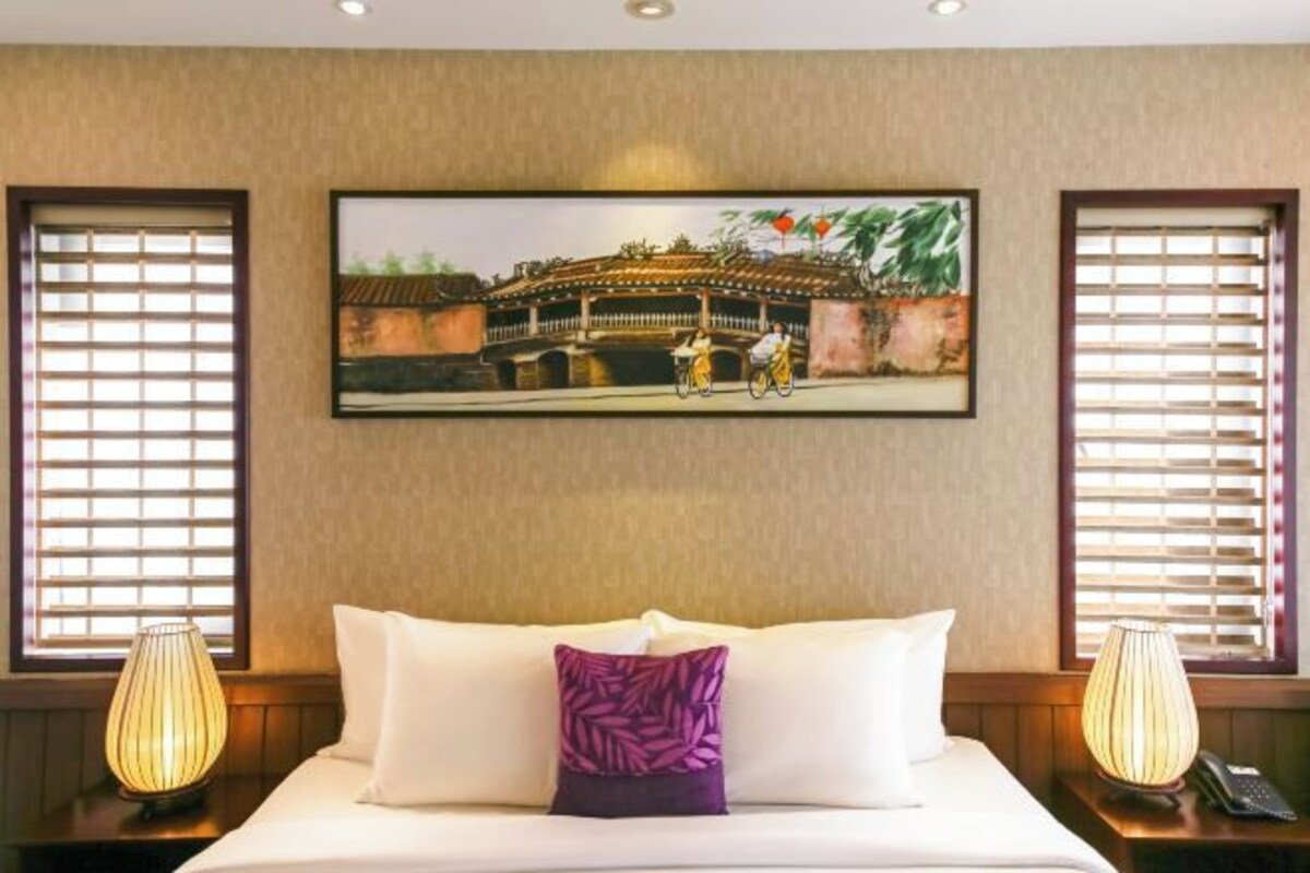 Una habitación de hotel con una cama, dos lámparas de noche, un cuadro en la pared y ventanas con persianas.
