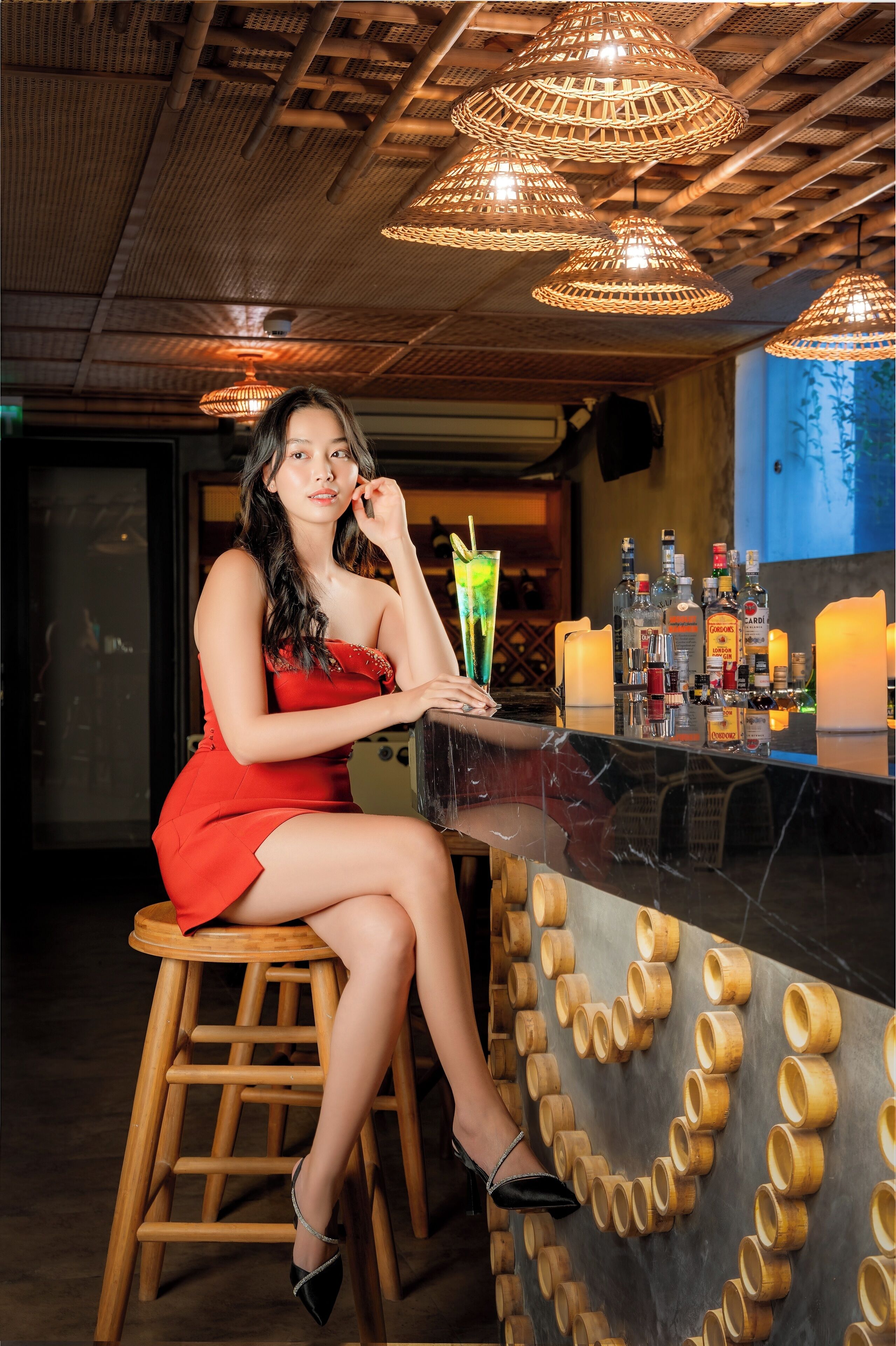 A person seated at a bar with a drink in hand, wooden stools, and a wall with cylindrical decorations.