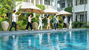 Zona piscina con persone che camminano sotto gli ombrelloni, una piscina cristallina e un edificio con balconi e vegetazione.