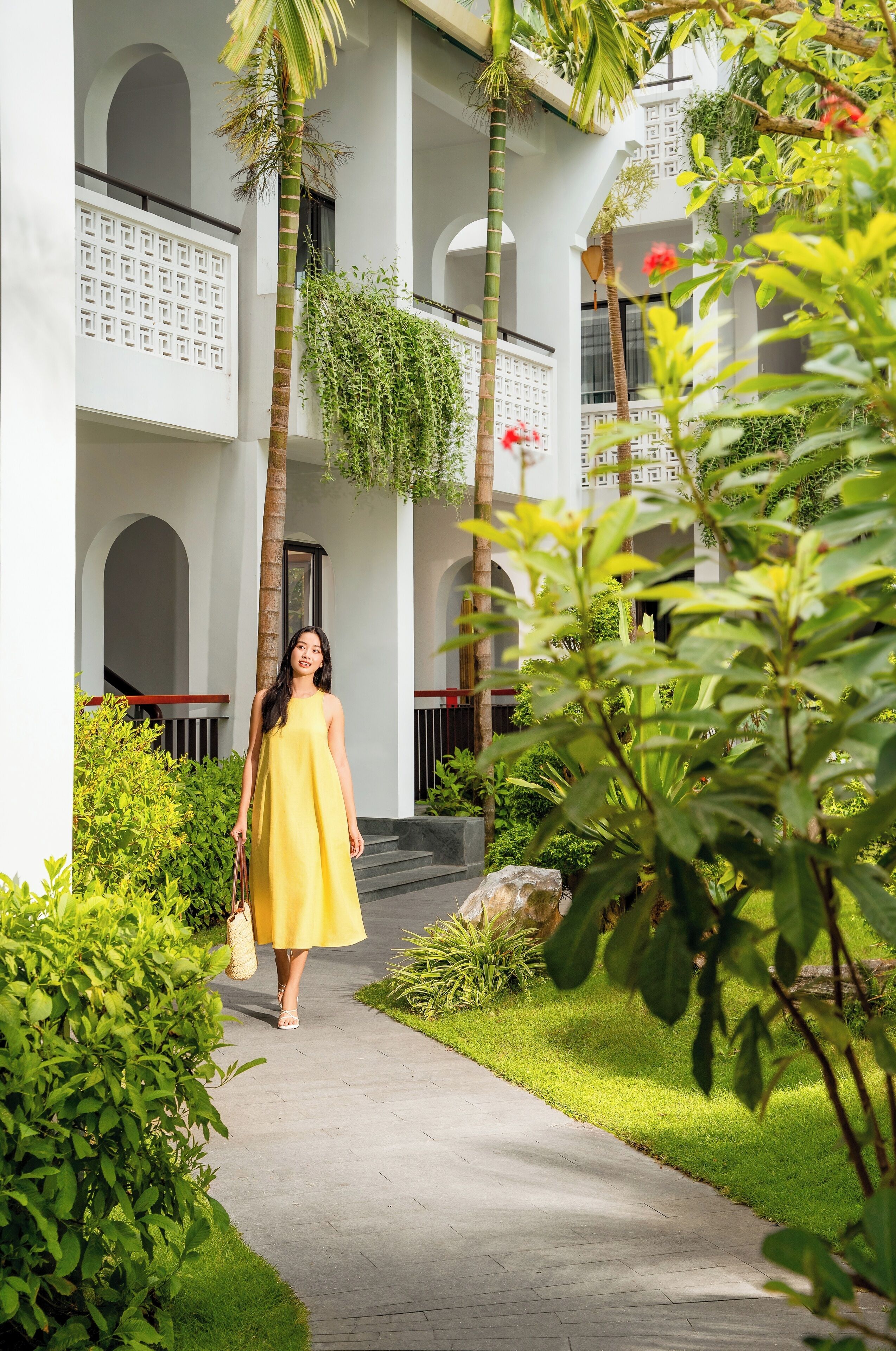 A woman in a yellow dress walking on a pathway surrounded by greenery and a white building with arched windows and a geometric design.