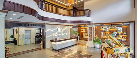 A hotel lobby with a reception desk, a piano, a staircase, and a decorative floor pattern.
