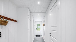 Un couloir aux murs et aux portes blancs, une étagère en bois avec un panier, et une vue sur un paysage verdoyant par une porte vitrée au fond.