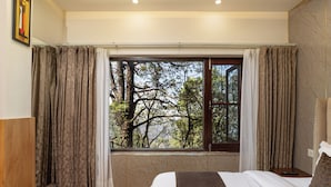 A bedroom with a large window, a bed with a brown quilt, and a nightstand with a remote control. - The Pine Retreat - Mall Road Dalhousie (Dalhousie)