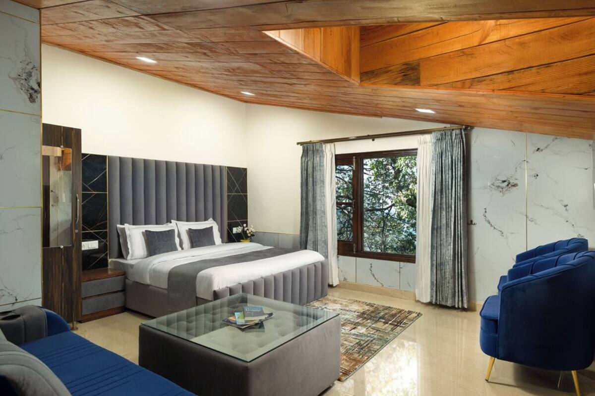 A modern bedroom with a large bed, a glass coffee table, a blue armchair, and a view of trees through the window.