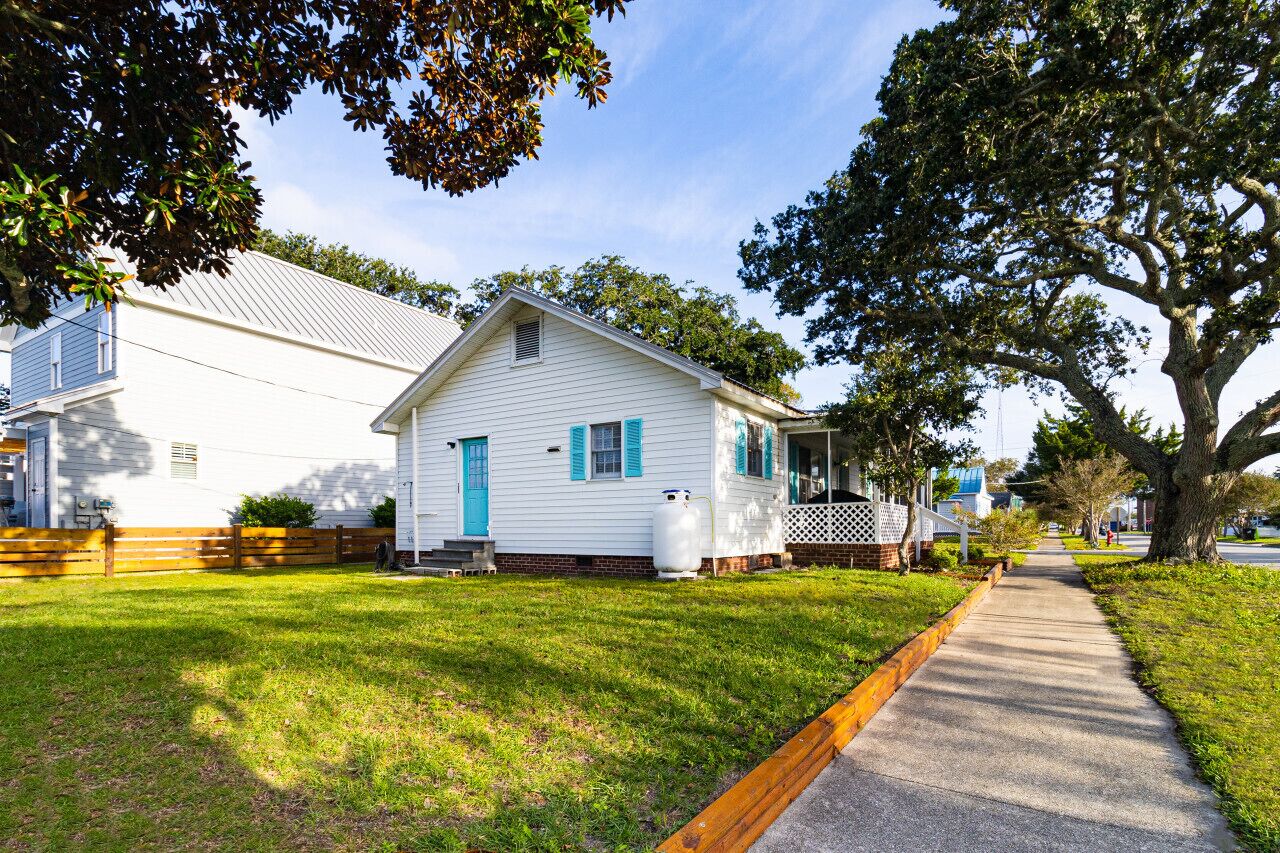 A white house with blue shutters, a front porch, and a large tree in front.
