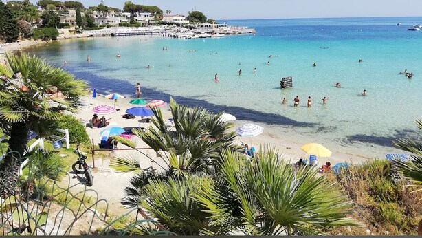 Una playa con agua turquesa cristalina, gente nadando y tomando el sol, y una hilera de edificios al fondo.