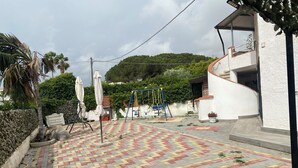 Un espacio al aire libre pavimentado con un parque infantil, que incluye columpios y un tobogán, rodeado de follaje y un edificio blanco.