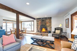 A spacious living room with a stone fireplace, wooden beams, a dining area, and a view of the outdoors.