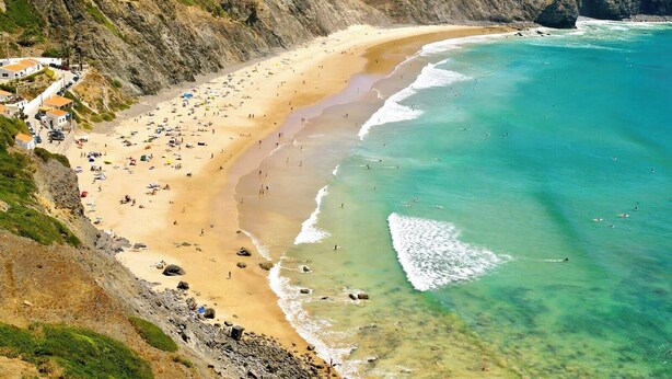 A coastal beach with a rocky cliff, sandy shore, and clear turquoise waters.