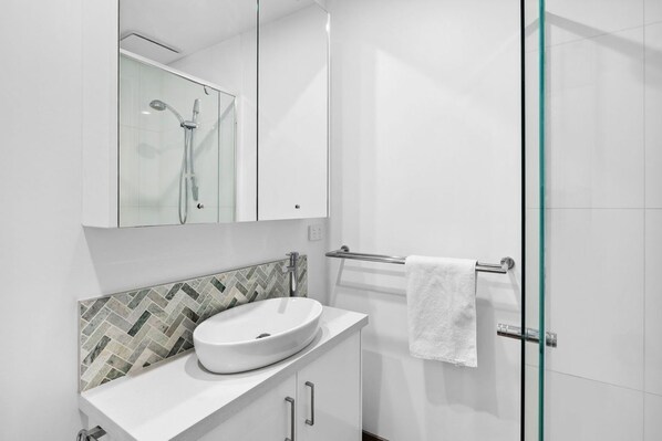 A modern bathroom with a glass shower enclosure, a white sink, and a herringbone-patterned tile backsplash.
