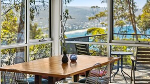 A dining area with a wooden table and metal chairs, large windows with a view of a beach and mountains, and a small round table.