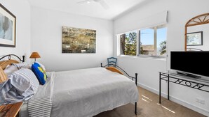 A bedroom with a bed, a television on a stand, a ceiling fan, a window with a view, and artwork on the wall.
