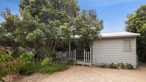 A small, single-story cabin with a porch, surrounded by dense vegetation and trees. - Coastwalk (Aireys Inlet)