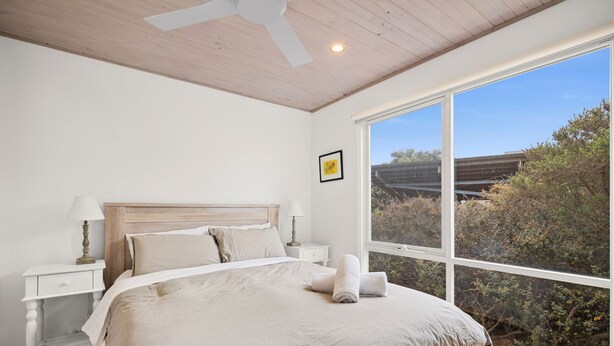 A bedroom with a large bed, two bedside tables, a ceiling fan, and a window with a view of trees and a building. - Coastwalk (Aireys Inlet)