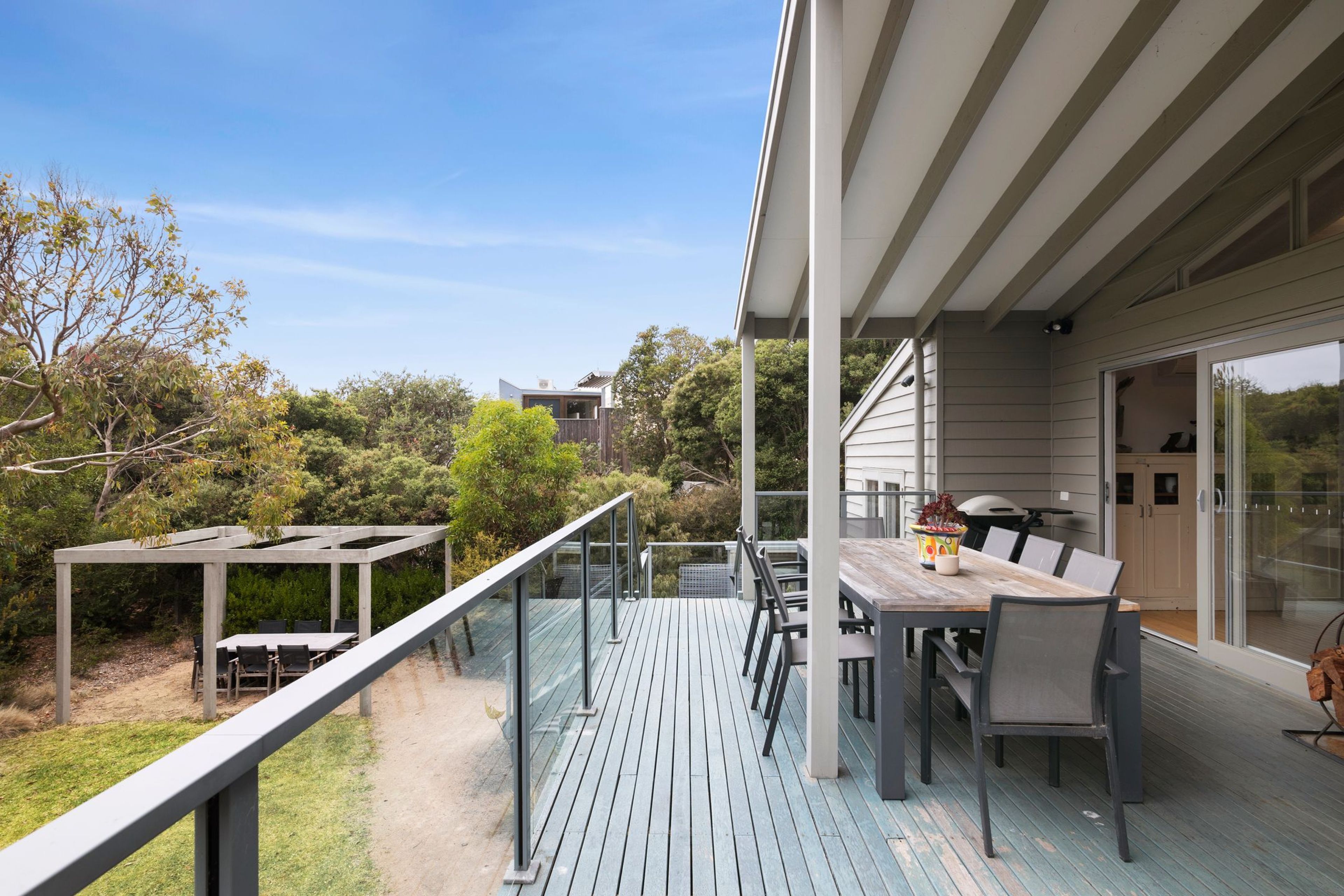 A modern outdoor deck with a dining area, surrounded by trees and a clear sky.