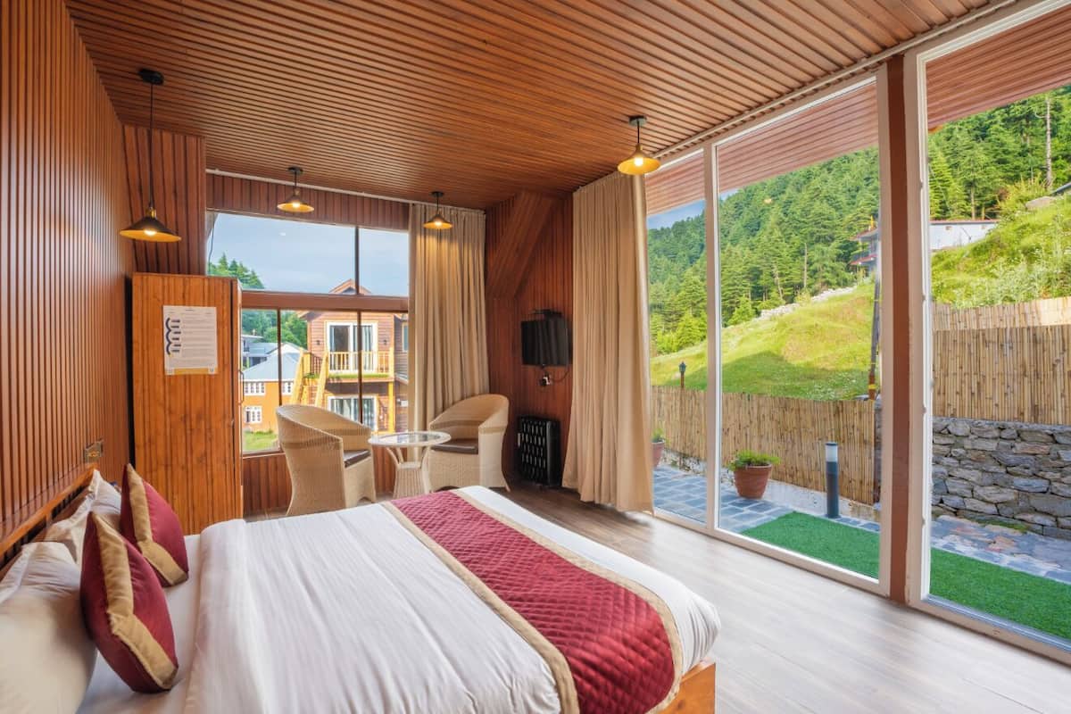 A spacious bedroom with a large bed, a wooden ceiling, and a sliding glass door leading to a balcony with a view of a green landscape.
