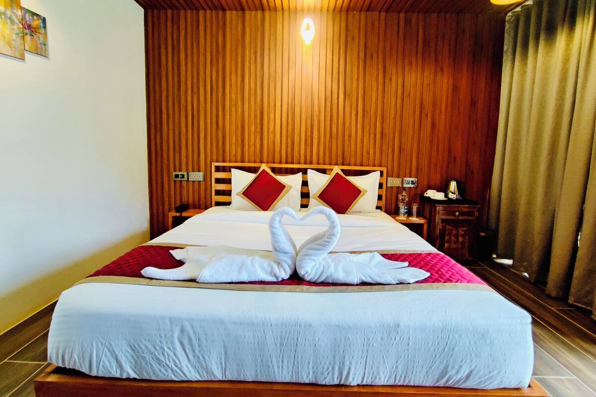 A hotel room with a wooden paneled ceiling, a bed with white bedding, red and white pillows, and a heart-shaped towel arrangement on the bed.