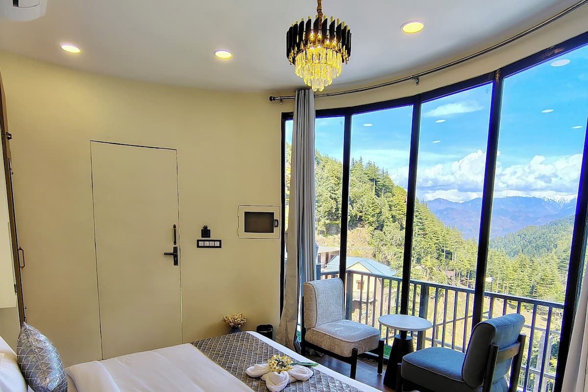 A hotel room with a large bed, a chandelier, a balcony with a table and chairs, and a view of mountains.