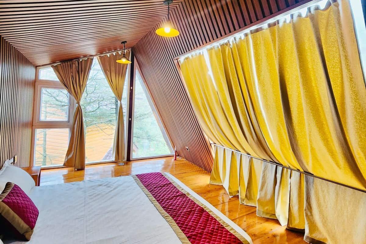 A room with a sloped ceiling, wooden flooring, a bed with a red accent, and large windows with yellow curtains.