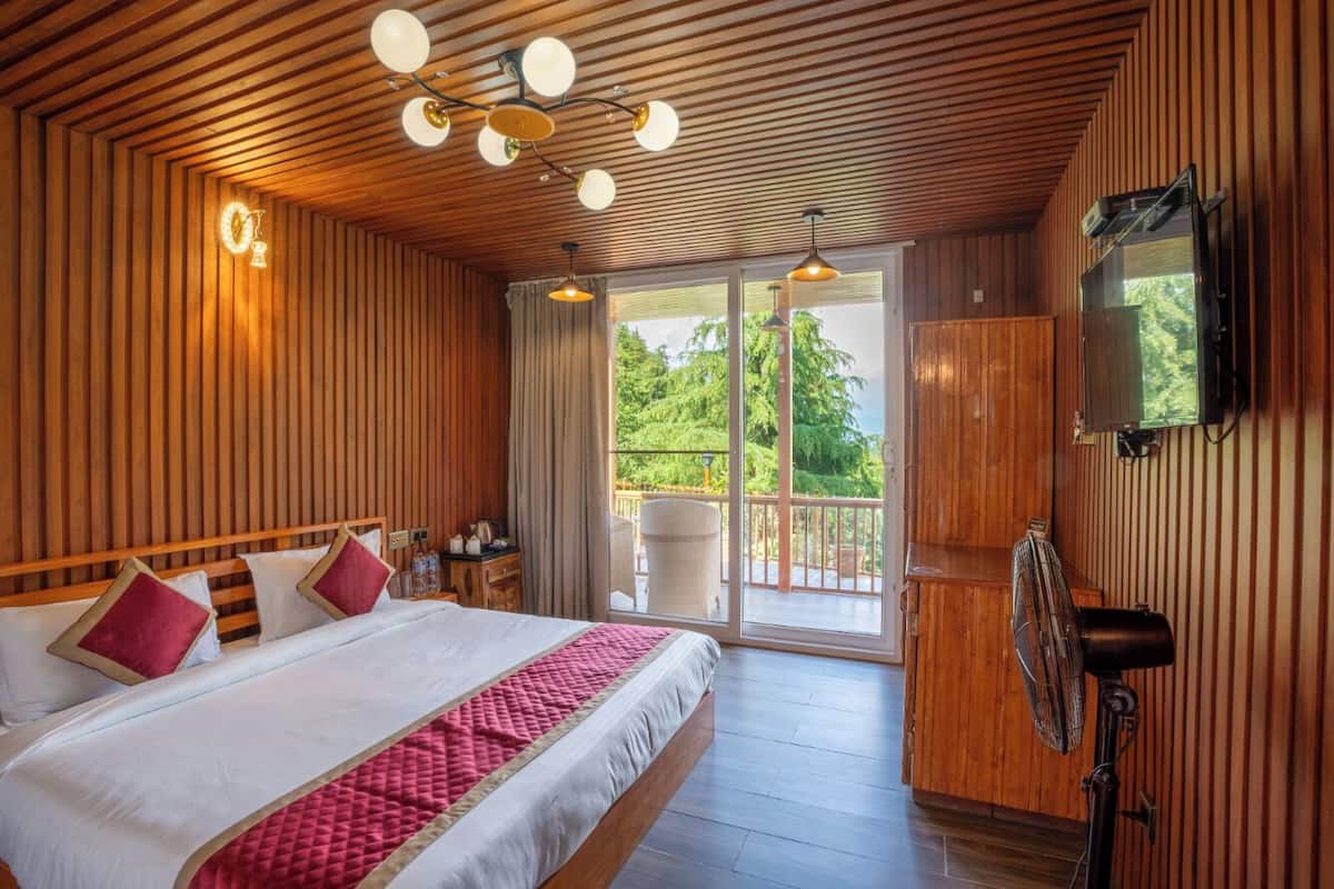 A spacious bedroom with a large bed, wooden paneling, a ceiling fan, and a balcony with a view of greenery.
