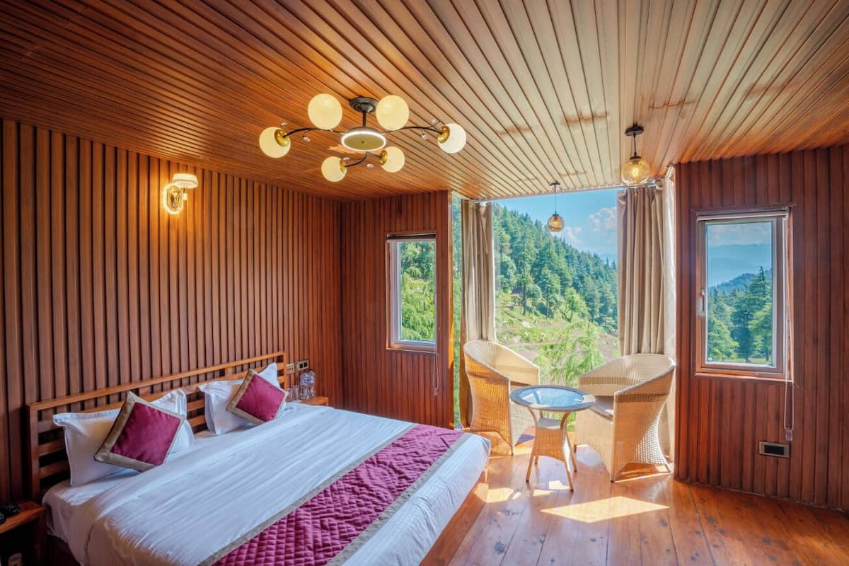 A wooden-paneled room with a large bed, a table with chairs, and a view of a forested landscape through windows.