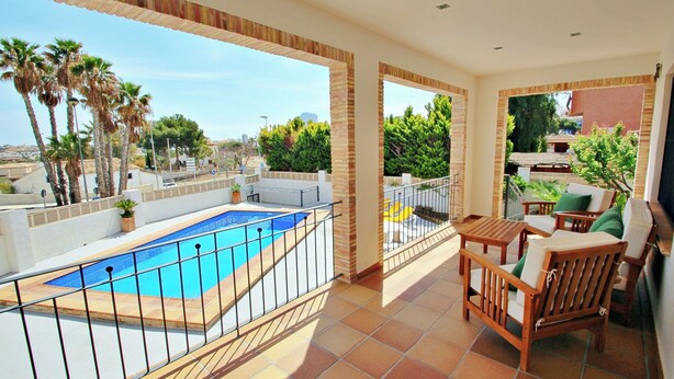 A balcony with a swimming pool, wooden furniture, and a view of palm trees and buildings.