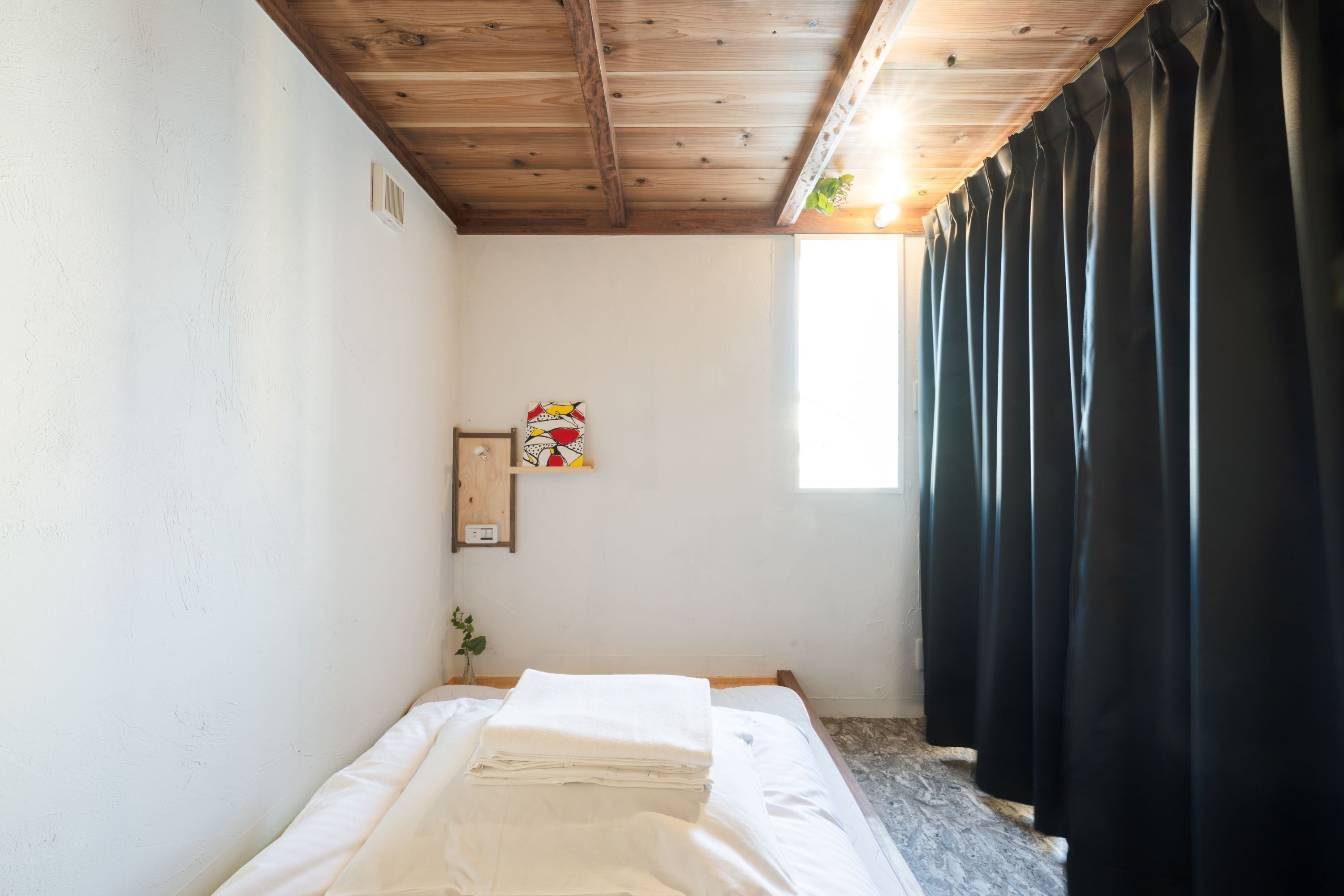A small, clean room with a wooden ceiling, a bed with white linens, a small shelf with a picture, and a window with black curtains.