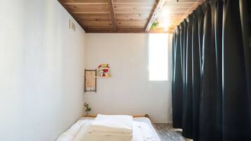 A small, clean room with a wooden ceiling, a bed with white linens, a small shelf with a picture, and a window with black curtains.