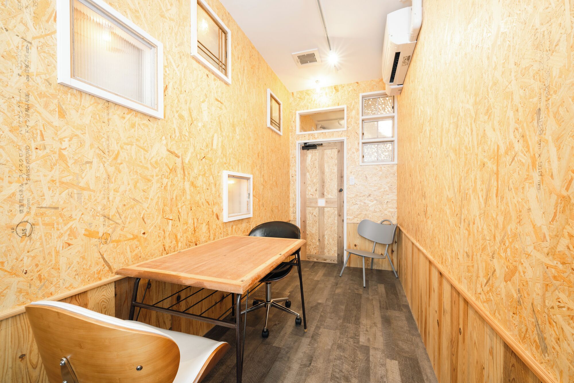 A narrow room with wooden walls, a wooden table, a chair, and a small desk with a chair. There are two windows and a door.