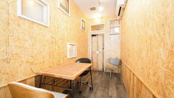 A narrow room with wooden walls, a wooden table, a chair, and a small desk with a chair. There are two windows and a door.